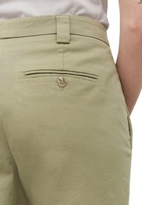 Pantalon kaki avec une poche passepoilée boutonnée à l’arrière, porté par une personne en chemise blanche.