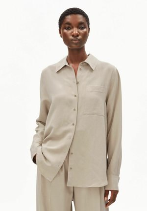 Vrouw draagt een losse beige overhemd met borstzak en bijpassende wijd uitlopende broek, staand tegen een effen witte achtergrond.