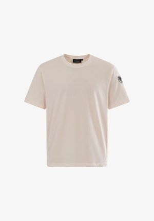 T-shirt beige à manches courtes et col rond, avec un petit patch noir et blanc sur la manche gauche et une étiquette de marque à l'intérieur du col.