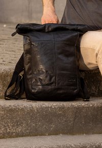 Crna kožna ruksak s preklopom i vidljivim šavovima, podesivim crnim trakama i suptilnom oznakom brenda sprijeda. Teksturirana završna obrada.