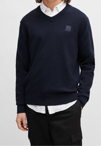 Tmavě modrý svetr s výstřihem do V z měkkého pleteného materiálu, s nenápadnou nášivkou a žebrovanými manžetami a lemem, nošený přes bílou košili s límečkem.