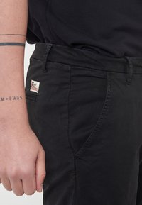 Pantaloni neri in tessuto liscio, con una tasca laterale, cuciture Sottili e un'etichetta con il marchio "Roy" posizionata vicino all'elastico in vita.