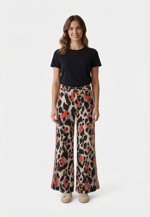 Donna in piedi, indossa una maglietta nera a maniche corte e pantaloni a gamba larga con motivo leopardato rosso, nero e beige, scarpe beige.