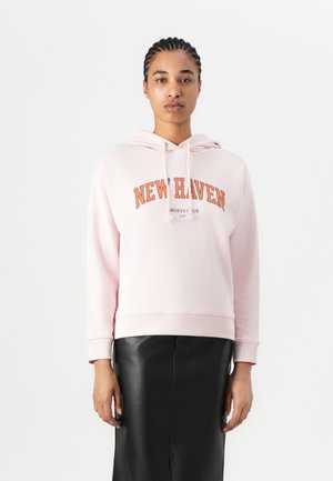Pink hoodie with "NEW HAVEN" printed in orange. Features a kangaroo pocket and drawstrings. Paired with a black leather skirt.