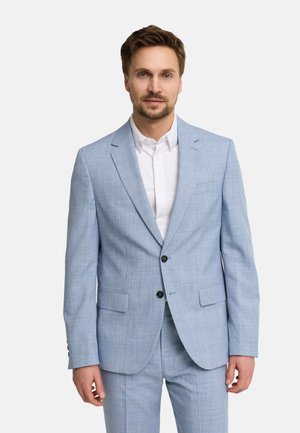 Man draagt lichtblauw pak en wit overhemd, staat voor een effen witte achtergrond, kijkt vooruit.