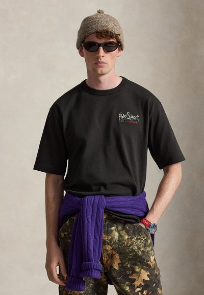 Νέος άντρας που φοράει μαύρο t-shirt Polo Sport, παντελόνι καμουφλάζ, μπεζ πλεκτό καπέλο, μαύρα γυαλιά ηλίου και μωβ πουλόβερ δεμένο γύρω από τη μέση του.