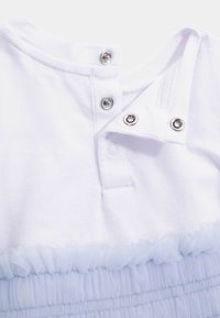 Valkoinen puuvillainen toppi, jossa on pyöreä pääntie, painonappikiinnitys ja alareunassa vaaleansininen röyhelöinen tylli.