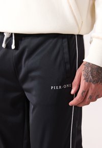 Black track pants made of smooth material featuring white side stripes and a logo reading "PIER·ONE" on the front near the pocket.