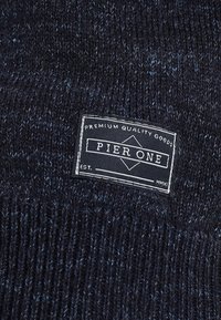 Tissu en tricot de pull en bleu foncé avec des flocons plus clairs. Présente une étiquette en tissu noir et blanc indiquant "Pier One" et "Produits de qualité supérieure."