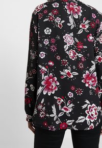 Blouse en tissu noir ornée de motifs floraux colorés en rouge, rose et blanc, avec des manches longues et un col rond.