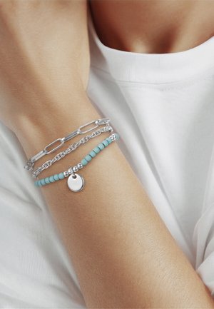 Pols met drie armbanden: zilveren schakels, zilveren ketting en turkooizen kralen met een ronde zilveren hanger, gedragen door een persoon met een wit overhemd.