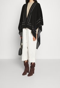 Svart poncho med grå ränder, lager-på-lager över en beige stickad topp. Kombinerad med vita byxor och bruna mockahöga stövlar. Svart handväska i handen.