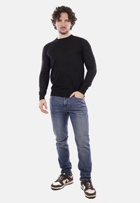 Camicia nera a maniche lunghe in tessuto liscio, abbinata a jeans in denim azzurro chiaro. Sneakers marroni e bianche con lacci. Design complessivo semplice.