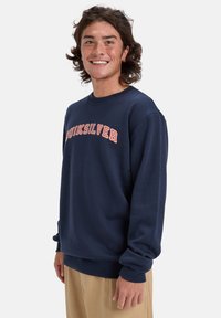 Námornícky modrý sveter vyrobený z mäkkého materiálu, s rebrovanými manžetami a pásom, na prednej strane s textom "Quiksilver" v svetlo oranžovej farbe v oblúku.