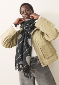 Black and gray patterned scarf with heart shapes and fringe edges, worn with a beige jacket and white top, displayed against a light background.
