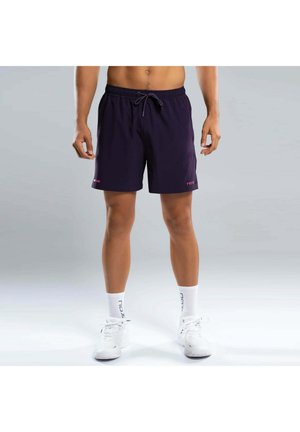 Uomo con pantaloncini sportivi neri con coulisse, calzini bianchi e scarpe da ginnastica bianche in piedi su uno sfondo grigio chiaro.