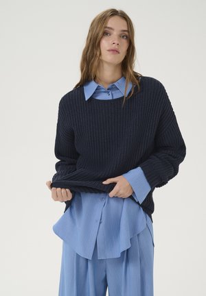 Maglione lavorato a maglia blu navy sopra una camicia colletto a righe verticali azzurro chiaro. Il maglione ha una vestibilità ampia e una texture a costine.