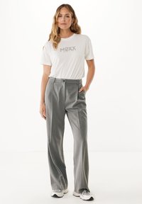 T-shirt bianca con grafica "MAXX", abbinata a pantaloni grigi plissettati. I pantaloni hanno una chiusura con bottone e tasche, con un design a gamba dritta.