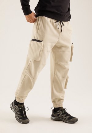 Homme portant un pantalon cargo beige avec des poches avant et latérales et des chaussures de sport noires, sur fond uni.