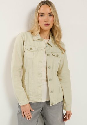 Mujer rubia que lleva una chaqueta beige abotonada sobre una camisa clara y pantalones grises, de pie frente a una pared lisa y clara.