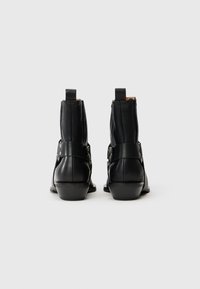 Botas de tobillo de cuero negro con un acabado suave, que cuentan con una correa con botones de metal y un tacón bajo y grueso. Asas resistentes en la parte posterior.