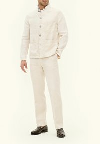 Giacca in lino beige chiaro con colletto alzato, tasche frontali e chiusura a bottoni, abbinata a pantaloni chiari coordinati e mocassini neri.