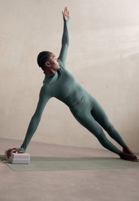 Weibliche Figur in einem grünen, eng anliegenden Langarmshirt und Leggings, die einen Side Plank auf einer Yogamatte ausführt, mit einem Yoga-Block unter einer Hand.
