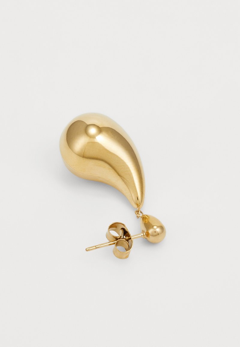 Clou d'oreille unique en forme de goutte dorée avec un petit pendentif en forme de goutte suspendu en dessous sur fond blanc.