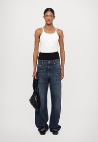 Top bianco senza maniche, jeans in denim a vita alta di un lavaggio blu scuro, vestibilità ampia, dettagli di cucitura minimi, abbinati a una borsa nera.