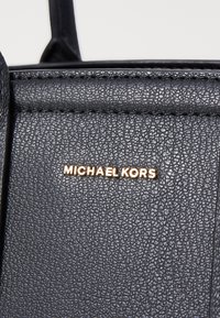 Mustat tekstuuriset nahkapinnat, joissa on näkyvä rakenne; korostaa kullankeltaista logoa, jossa lukee "MICHAEL KORS."