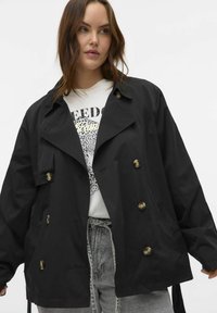 Sort trenchcoat med en afslappet pasform, dobbeltknappede bronzeknapper, stor krave og tekstureret stof over en hvid grafisk t-shirt.