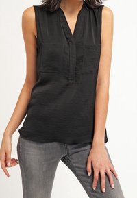 Top negro sin mangas con escote en V, dos bolsillos delanteros, textura suave y un corte relajado, combinado con jeans de mezclilla grises.