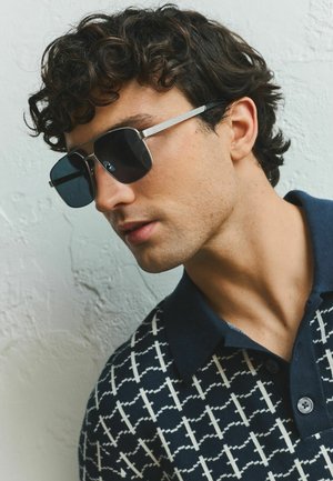 Junger Mann mit dunklem lockigem Haar, der große silbergerahmte Sonnenbrillen und ein marineblaues Hemd mit Muster und Kragen trägt, vor einer weißen strukturierten Wand.