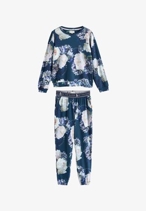 Vrouw staat blootsvoets binnen, gekleed in een blauw loungewear set met bloemenprint, drinkt water uit een glas, met één hand in de zak dichtbij het raamlicht.
