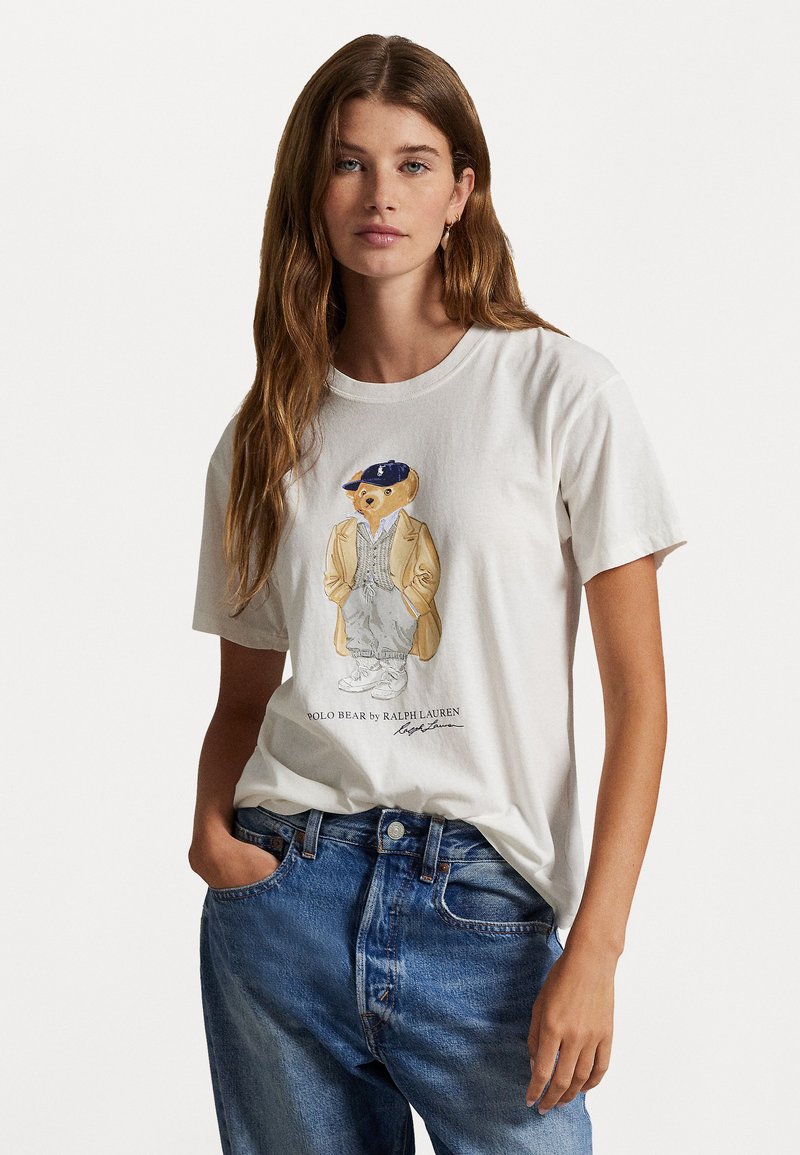 White cotton T-shirt featuring a cartoon bear graphic in a blazer and hat, with blue jeans. Simple crew neckline and relaxed fit.