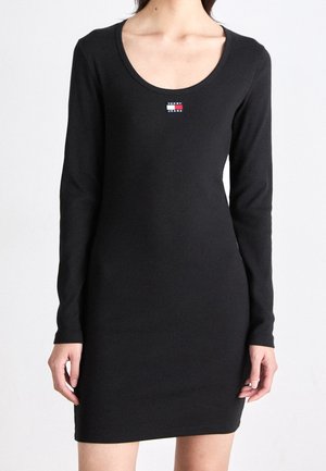 Kobieta w dopasowanej czarnej sukience z długim rękawem, z zaokrąglonym dekoltem oraz małym logo Tommy Jeans na piersi.