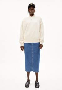 Licht beige oversized sweatshirt met ribbelkraag, gecombineerd met een lange blauwe denimrok. Zwarte instappers en een zwarte pet met een kleurrijk ontwerp.