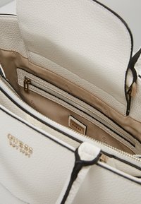 Valkoinen nahkalaukku, jossa on teksturoidun pinnan, beige vuori, sisempi vetoketjupussi ja musta korostusreuna. Kullanvärinen logo näkyvissä.