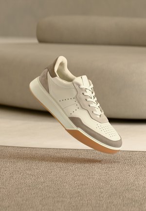 Witte en beige sneaker met witte veters en een gumzolen, zwevend boven een beige tapijt voor een bijpassende beige bank.