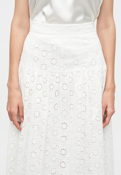 Γυναίκα με λευκή φούστα από δαντέλα με λουλούδια και σατέν τοπ, τα χέρια χαλαρά στα πλευρά.
