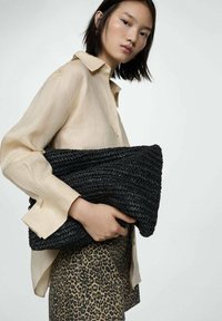Crna pletena clutch torbica koju drži model, s pravokutnim oblikom i teksturiranom površinom, u kombinaciji s bež košuljom i suknjom s leopard uzorkom.
