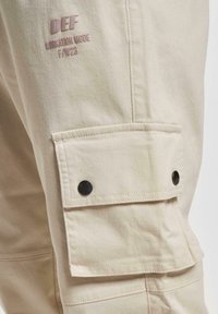 Beige cargo pants with a side pocket and black snap closure. Features brand logo in light pink, textured cotton fabric, and clean stitching.