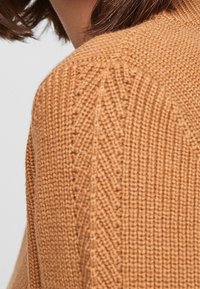 Gebreide trui in warm beige met een textuurpatroon, met ribbel details en een close-up van het schoudergedeelte.