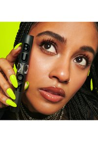 Czarna tubka z etykietą "BUXOM" i srebrnymi akcentami, trzymana w dłoni z jaskrawozielonymi paznokciami. Tło to jasny neonowy żółty. Nałożony przezroczysty błyszczyk do ust.