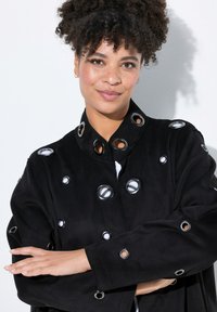 Chaqueta negra con ojales circulares, con un alto cuello y cierre de botones. Tela suave con un corte relajado y grandes acentos de ojal.