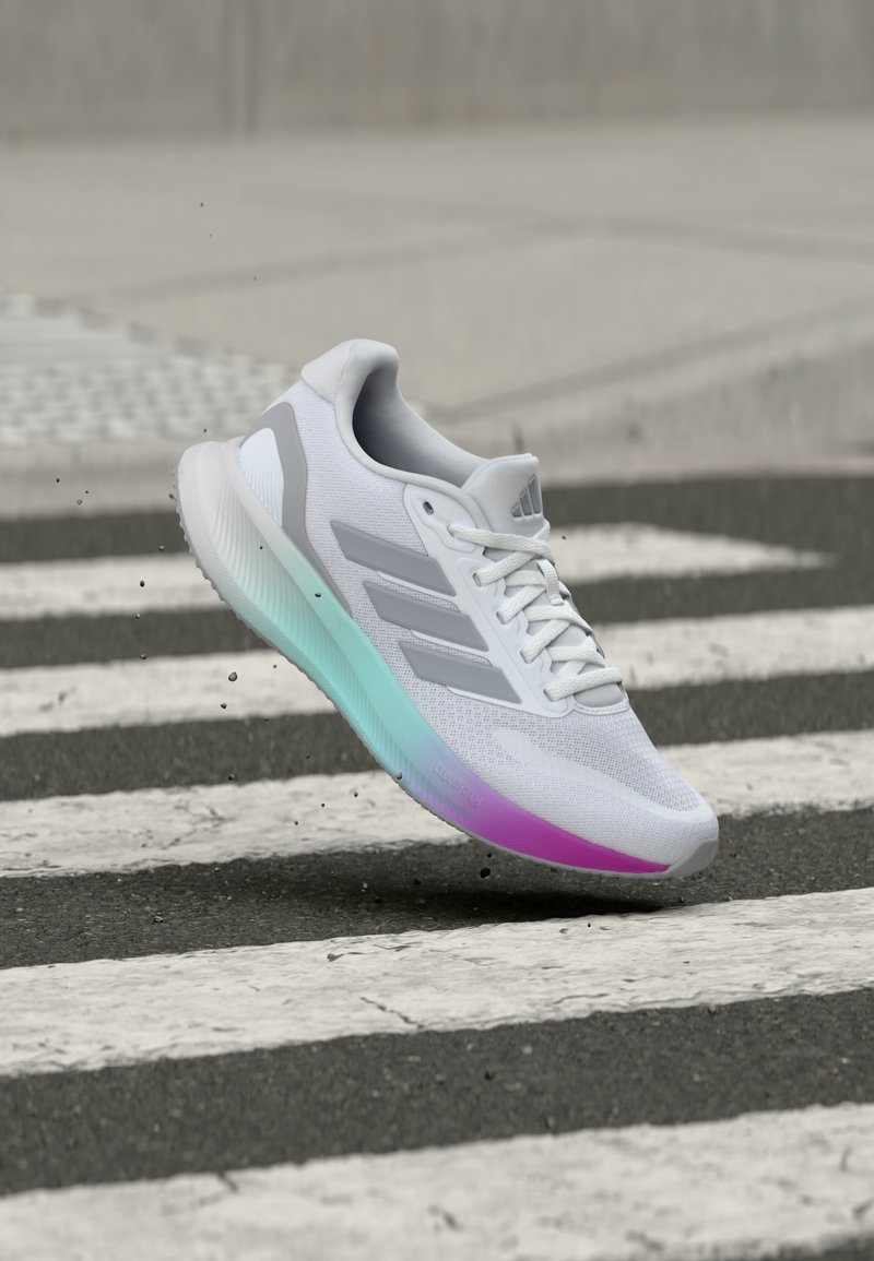 Baskets de sport blanches avec des rayures grises et une semelle dégradée flottant au-dessus d'un passage piéton noir et blanc, soulevant de petites particules de saleté.