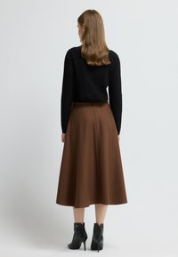 Falda marrón de longitud hasta la rodilla, en línea A, con tela texturizada, cierre de cremallera lateral, combinada con un suéter negro y botines de tacón negros.