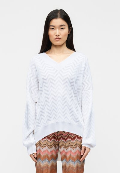Pull en maille léger blanc à col en V, présentant un motif en zigzag et des manches larges, assorti à un pantalon coloré à motifs.