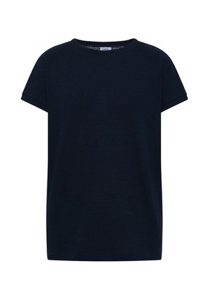 Femme portant un top noir à manches courtes, un jean court bleu clair, des baskets argentées, une casquette rose, tenant un petit sac marron dans une pièce aux tons neutres.