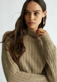 Maglione beige a trecce con collo alto, caratterizzato da scanalature testurizzate. La modella indossa orecchini a cerchio dorati e mostra lunghi capelli ondulati.